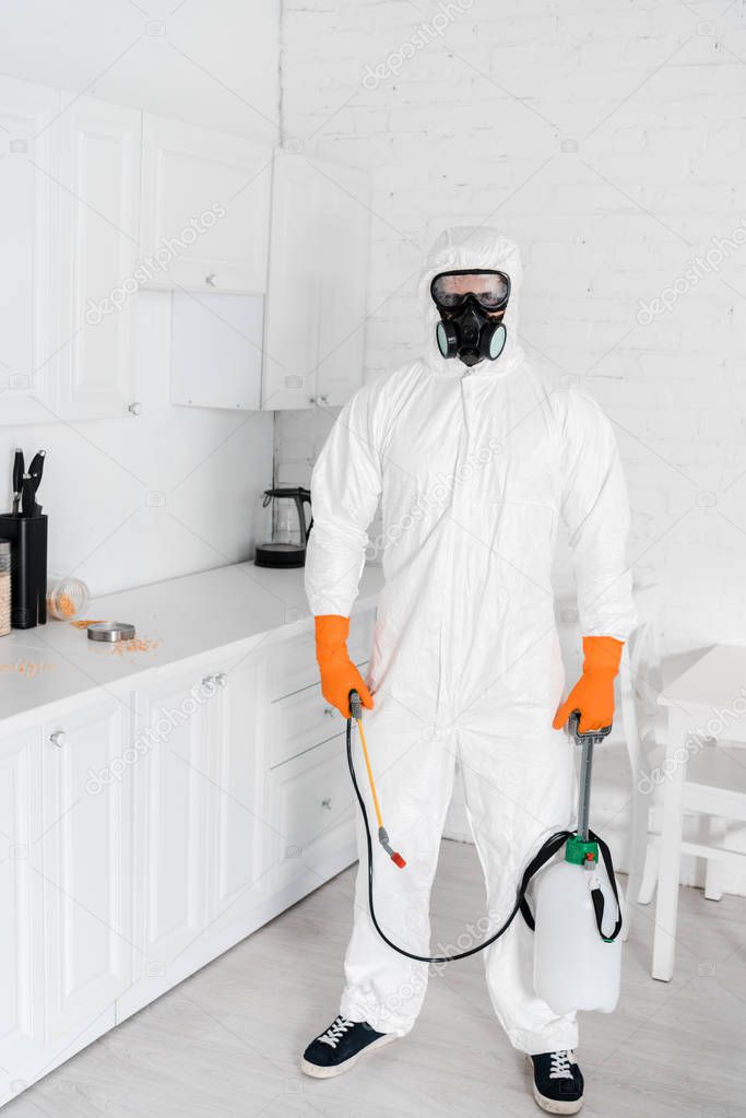 exterminator in protective mask and uniform holding toxic equipment while standing near kitchen cabinet 