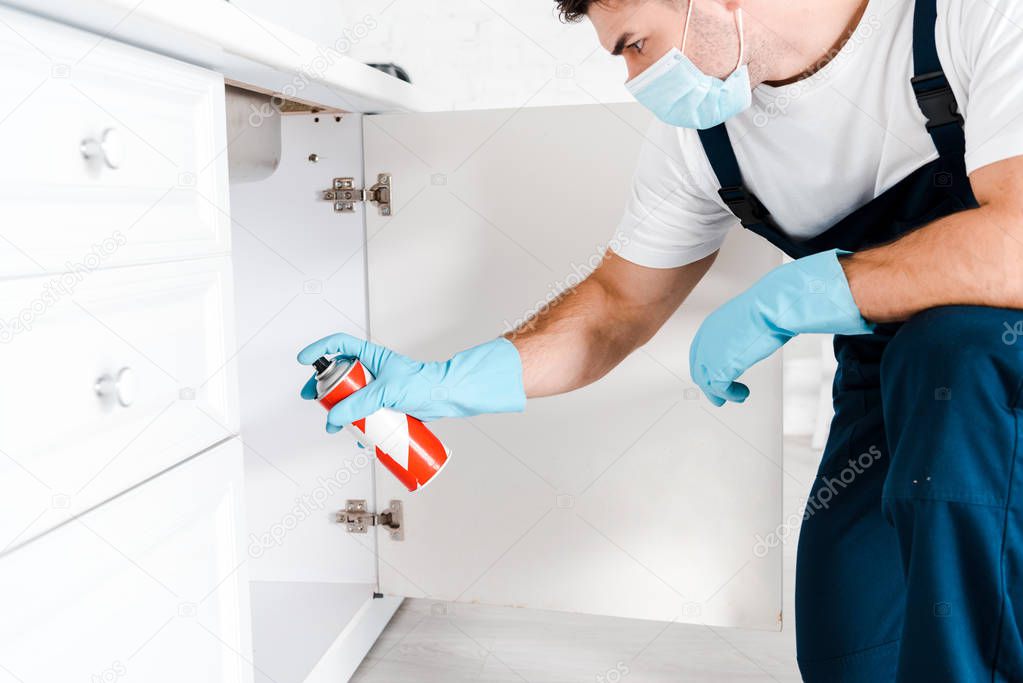 exterminator in protective mask holding spray can near kitchen cabinet 