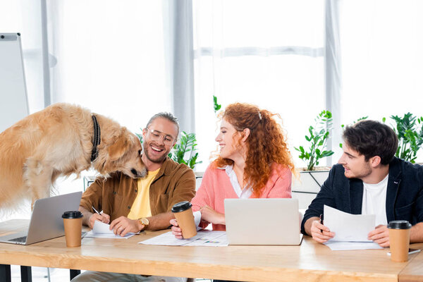 three friends smiling and looking at cute golden retriever in office 