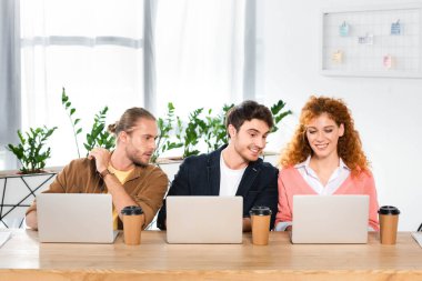 masada oturan ve ofiste dizüstü bilgisayar kullanan üç gülümseyen arkadaş 