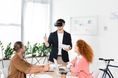 smiling businessman with virtual reality headset holding pen and papers and friends looking at him  clipart