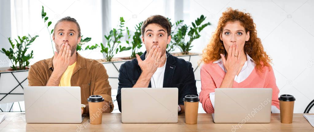 panoramic shot of three shocked friends sitting at table and obscuring faces 