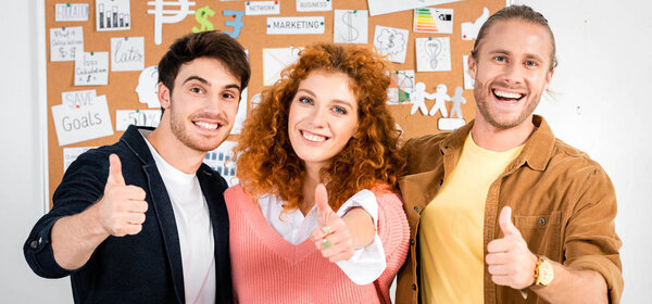 panoramic shot of three smiling friends showing thumbs up and looking at camera 