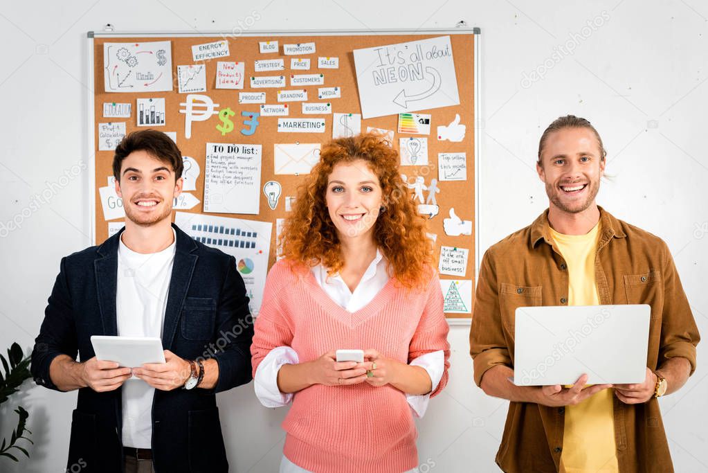 three smiling friends holding laptop, smartphone and digital tablet in office 