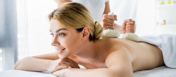 panoramic shot of masseur doing back massage with herbal balls to smiling woman in spa 