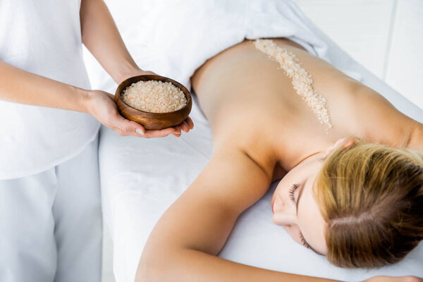 cropped view of masseur holding bowl with sea salt and woman lying on massage mat in spa 