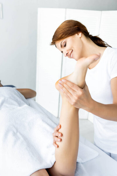 attractive and smiling masseur doing foot massage to woman in spa 