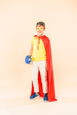full length view of boy in mask and red cloak with skateboard on pink clipart