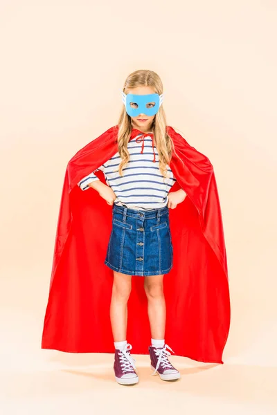 full length view of child in mask and hero cloak standing with hands on hips on pink