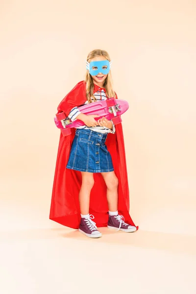 full length view of child in mask and hero cloak holding skateboard on pink