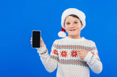 pohled na usmívající se dítě v Santa klobouku ukazující prstem na smartphone s prázdnou obrazovkou izolovanou na modré