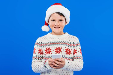 mavi izole akıllı telefon kullanarak santa şapka gülümseyen çocuk ön görünümü