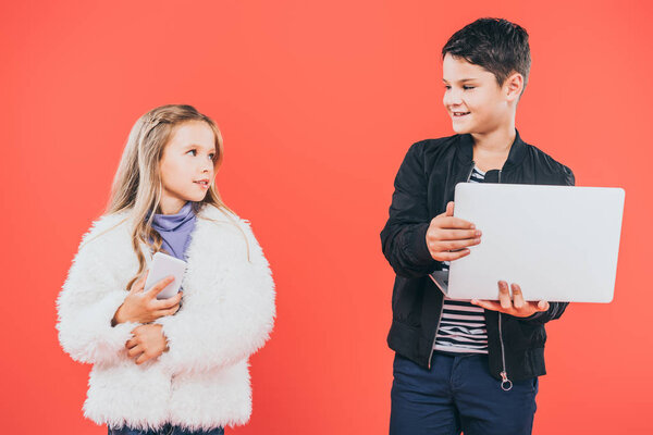 smiling kid with smartphone and laptop looking at each other isolated on red
