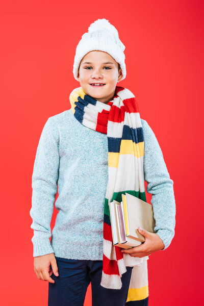 front view of smiling kid in hat and scarf holding books isolated on red