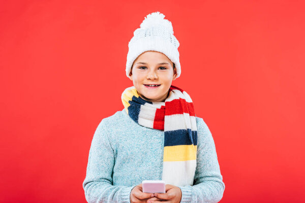 front view of smiling kid in hat and scarf using smartphone isolated on red