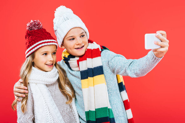 two smiling kids in winter outfits embracing and taking selfie isolated on red