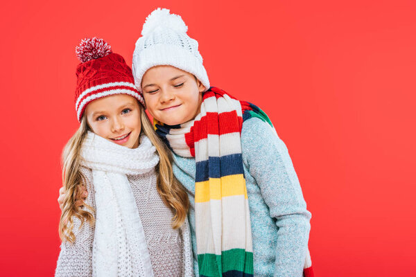 two smiling kids in hats and scarfs embracing isolated on red