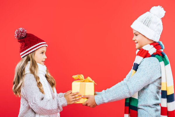 side view of kids in winter outfits holding present and looking at each other isolated on red