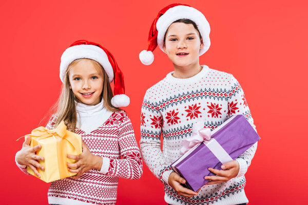 front view of two smiling kids in santa hats with presents isolated on red