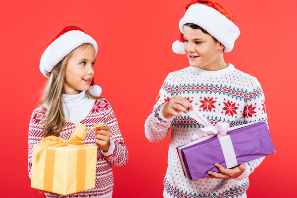 two smiling kids in santa hats with presents looking at each other isolated on red
