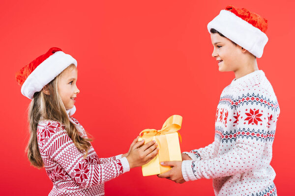 two smiling kids in santa hats with present looking at each other isolated on red