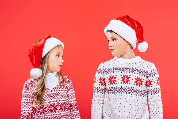 Dwa Zdziwione Dzieci Kapelusze Santa Patrząc Siebie Izolowane Czerwono — Zdjęcie stockowe
