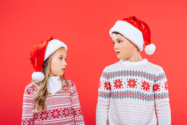 two surprised kids in santa hats looking at each other isolated on red