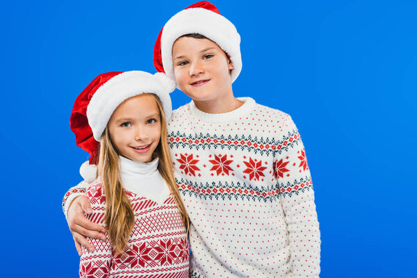 two smiling kids in sweaters and santa hats embracing isolated on blue