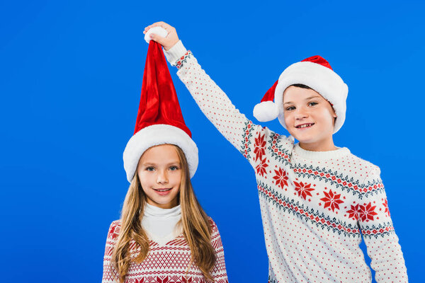 two smiling kids in sweaters and santa hats isolated on blue