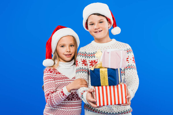 front view of two smiling kids in santa hats with gifts isolated on blue