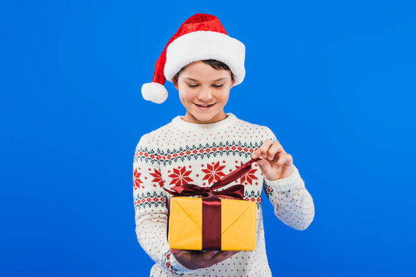 front view of kid in santa hat and sweater holding gift isolated on blue