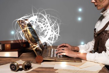 cropped view of woman typing on steampunk laptop with glowing digital illustration at table isolated on grey clipart