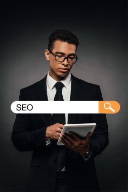 Gözlüklü Afro-Amerikan iş adamı karanlık arka planda dijital tablet kullanıyor ve arama çubuğu resimlerinde seo harfleri var.