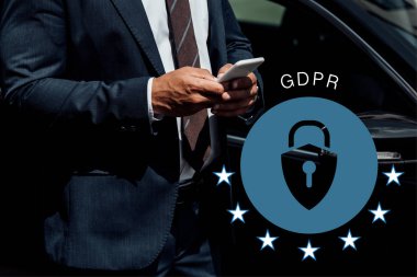 Afrika kökenli, takım elbiseli Amerikalı iş adamının güneşli bir günde arabanın yanında GDPR illüstrasyonlu akıllı telefon kullanması.