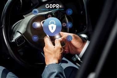 Afrika kökenli Amerikalı takım elbiseli iş adamının güneşli bir günde arabada GDPR illüstrasyonlu akıllı telefon kullanmasının kısmi görüntüsü