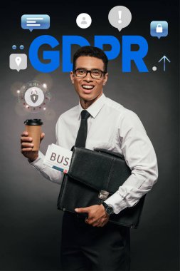 Mutlu Afro-Amerikan iş adamı elinde gazete, paket kahve ve koyu arkaplanda GDPR illüstrasyonlu deri çanta tutuyor.