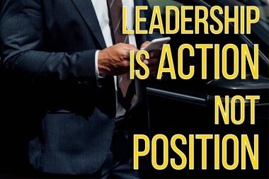 cropped view of african american businessman in suit using smartphone near car with leadership is action not position lettering clipart