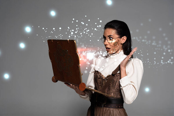 shocked steampunk woman in glasses using vintage laptop with glowing illustration isolated on grey