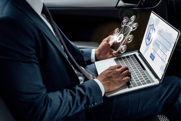 cropped view of african american businessman using laptop and smartphone in car with internet security illustration