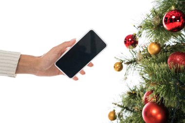 Kırpılmış bir görüntüde, beyaz üzerinde izole edilmiş noel ağacının yanında boş ekranlı akıllı telefon tutan bir kadın görülüyor. 
