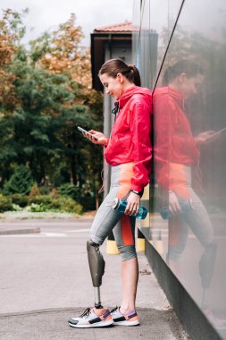 Kulaklıkla müzik dinleyen ve sokakta akıllı telefon kullanan engelli sporcu kadının yan görüntüsü