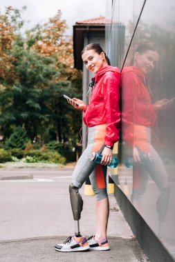 Spor şişesi ve akıllı telefonuyla gülümseyen engelli kadın tam boy görünümü