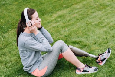 Çimlerin üzerinde oturan ve kulaklıkla müzik dinleyen engelli sporcu kadının yan görüntüsü