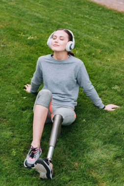Engelli sporcu kadın çimde oturuyor ve kulaklıkla müzik dinliyor.
