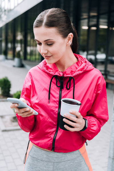 smiling sportswoman holding cup and using smartphone on street