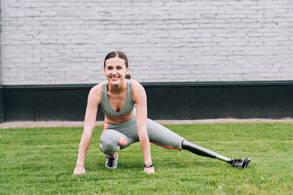 smiling disabled sportswoman with prosthesis stretching on grass