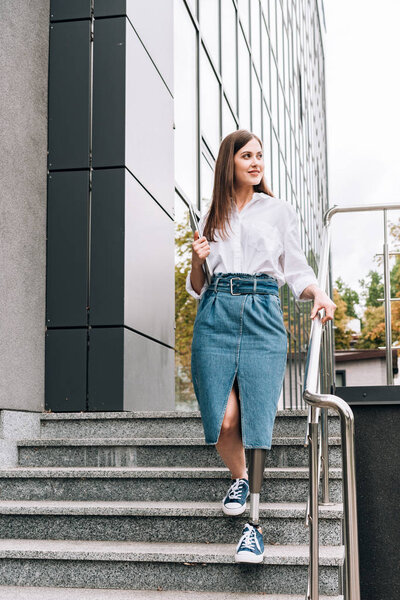 Вид в полный рост женщины-инвалида в джинсовой юбке, держащей ноутбук на улице
