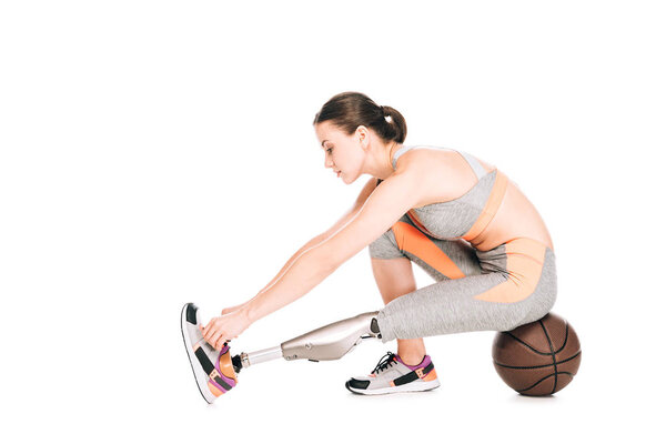 side view of disabled sportswoman with prosthesis sitting on basketball ball and tying shoelaces isolated on white
