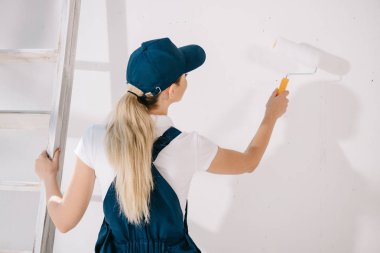 back view of young painter in uniform standing near white wall and holding paint roller clipart