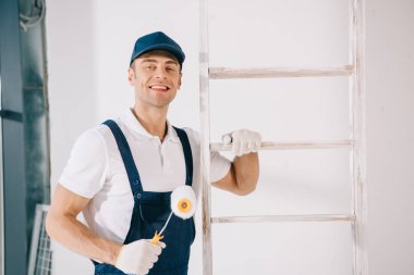 handsome young painter in uniform smiling at camera while standing near ladder and holding paint roller clipart